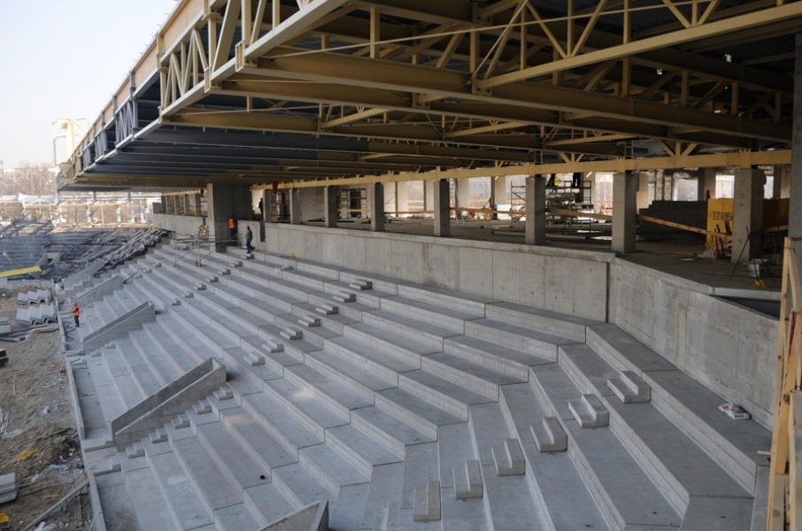 Betonowe trybuny w trakcie budowy stadionu, widoczne elementy konstrukcji stalowej dachu i pracujący robotnicy w oddali.