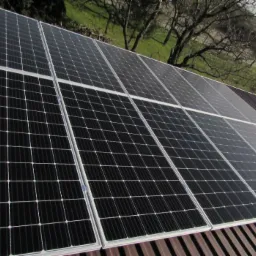 Panel słoneczny na dachu z widoczną teksturą blachy i drzewami w tle, uchwycony pod lekkim kątem.