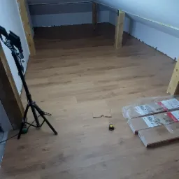 Montaż paneli podłogowych na poddaszu, widoczny statyw kamery, miarka i opakowania paneli Egger Pro.