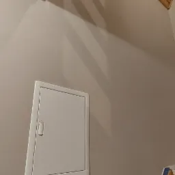 Beżowa ściana na poddaszu z drewnianym wykończeniem sufitu, białą klapą rewizyjną i brązowymi włącznikami światła.