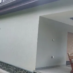 Świeżo otynkowana elewacja domu jednorodzinnego z widocznymi detalami wykończeniowymi, takimi jak okładzina kamienna przy fundamencie, okna z roletami zewnętrznymi i kamienne filary werandy.