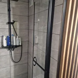 Nowoczesna kabina prysznicowa z czarnym osprzętem i szklaną ścianką, płytki imitujące marmur na ścianach, oraz dekoracyjny panel z drewnianych listew.