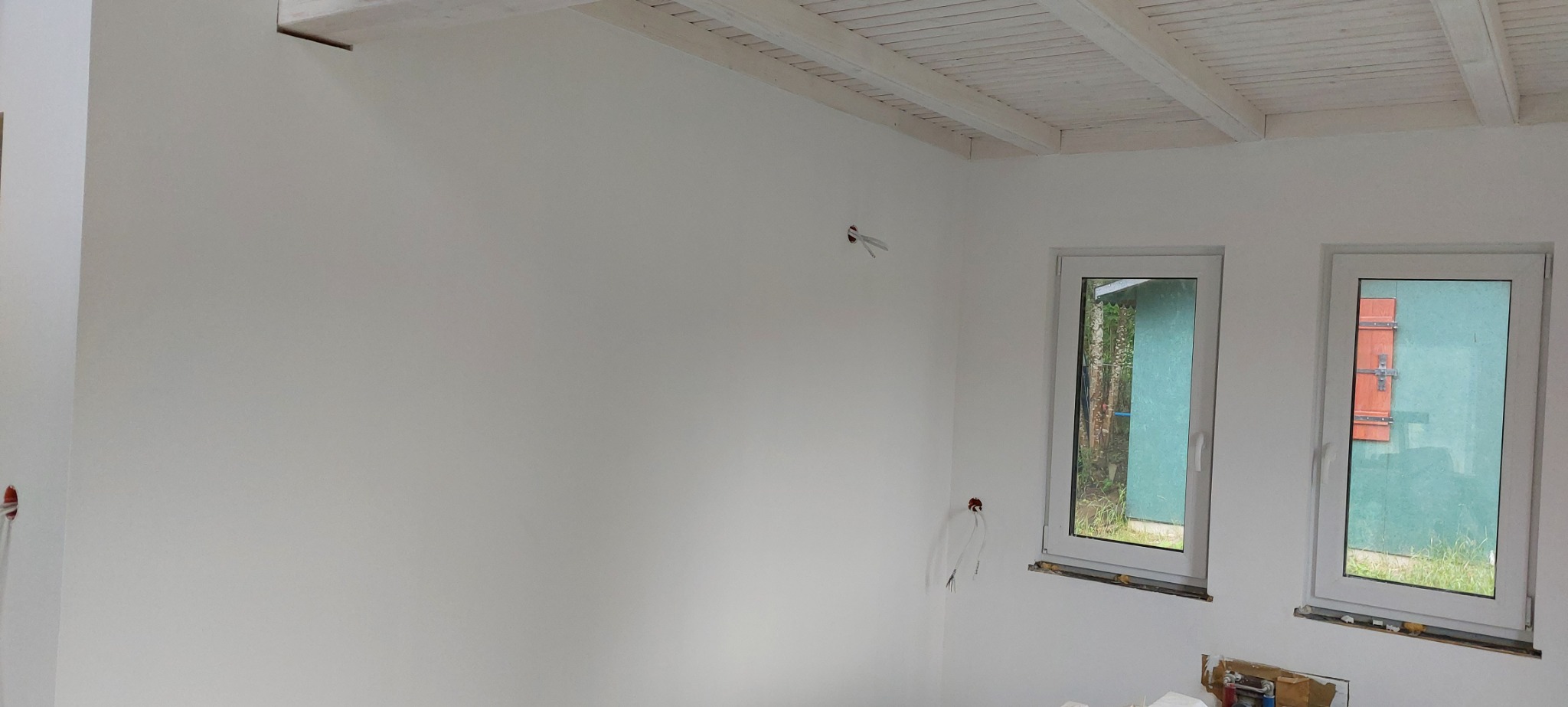 Wnętrze po malowaniu na biało, widoczne dwa okna z widokiem na zewnątrz, biały sufit z drewnianymi belkami, wystające kable elektryczne ze ściany.