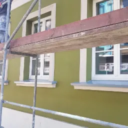 Pracownik na rusztowaniu maluje elewację budynku na oliwkowo-zielony kolor z białymi detalami wokół okien.