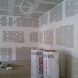 Wnętrze w trakcie ocieplania poddasza: widoczne płyty gipsowo-kartonowe na ścianach i suficie, nieotwarte rolki wełny mineralnej Climowool oraz zwisający kabel elektryczny.