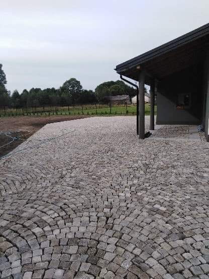 Ułożona kostka granitowa w kształcie wachlarza na podjeździe przed domem z szarą elewacją i drewnianym dachem, widoczne dwie kolumny podtrzymujące dach.