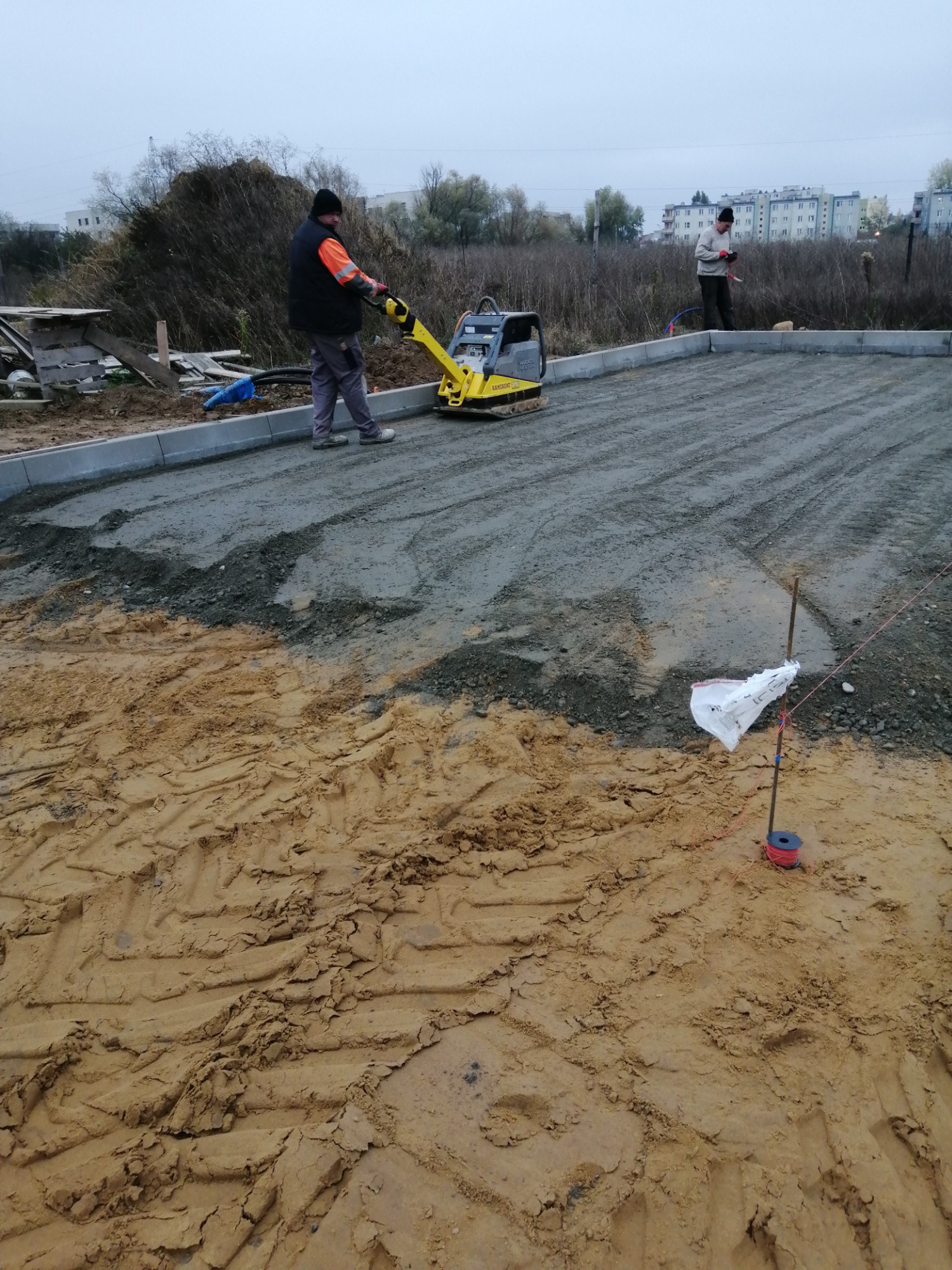 Prace przygotowawcze pod nawierzchnię: ubijanie podłoża zagęszczarką wibracyjną, widoczne obrzeża i wytyczone granice przyszłej powierzchni.