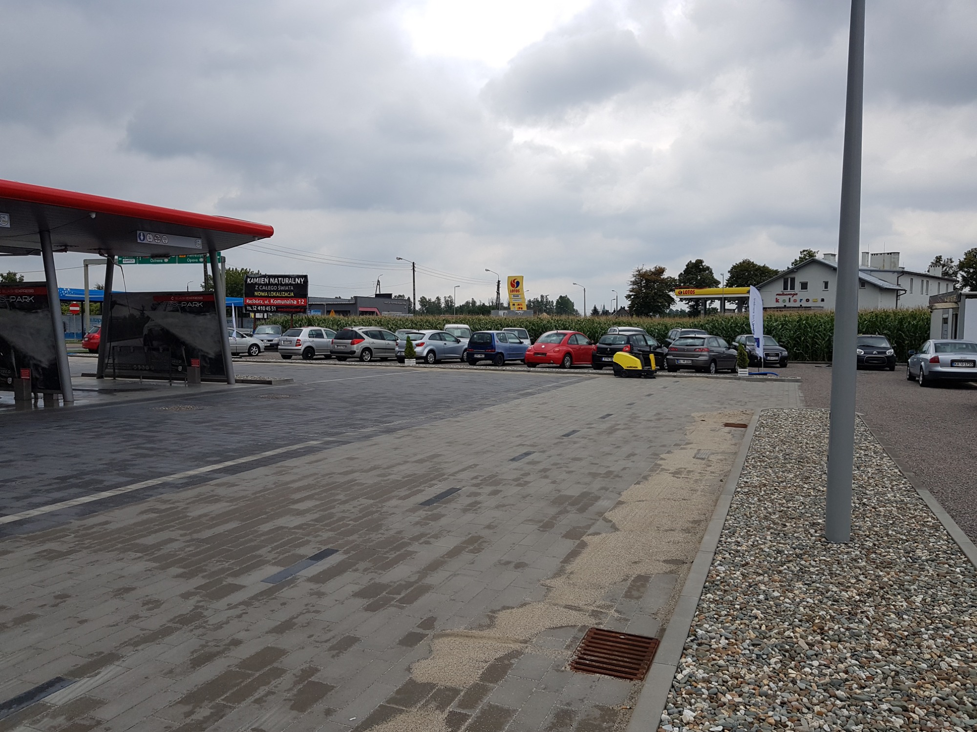 Szara kostka brukowa na parkingu przy stacji benzynowej i myjni samochodowej, widoczny odpływ liniowy i samochody w tle.