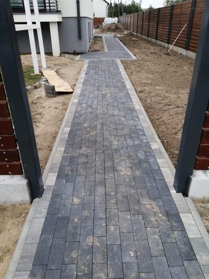 Ułożona ścieżka z ciemnoszarej kostki brukowej z jasnoszarą obwódką prowadząca wzdłuż ogrodzenia i budynku. Widoczne ślady piasku na powierzchni kostki i niezakończone prace w tle.