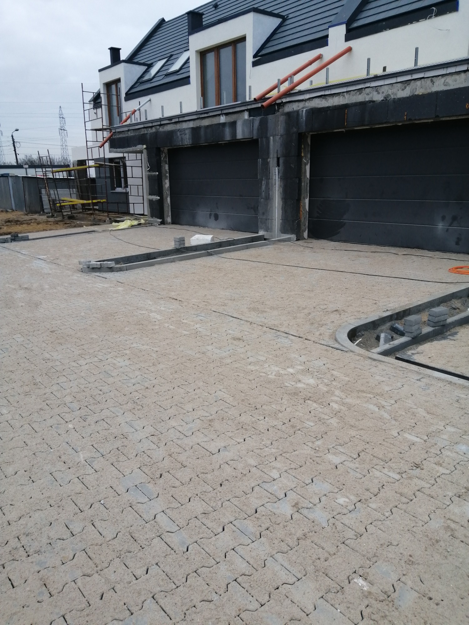 Układanie szarej kostki brukowej typu 'I' na podjeździe do garażu w trakcie realizacji, widoczne krawężniki i przygotowane podłoże, budynek w tle w trakcie wykończenia.