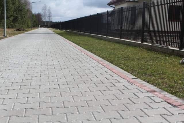 Długa, prosta ścieżka ułożona z szarej kostki brukowej typu 'I', obramowana czerwonymi kostkami krawężnikowymi, biegnąca wzdłuż czarnego, metalowego ogrodzenia i trawnika. W tle widoczny słup...