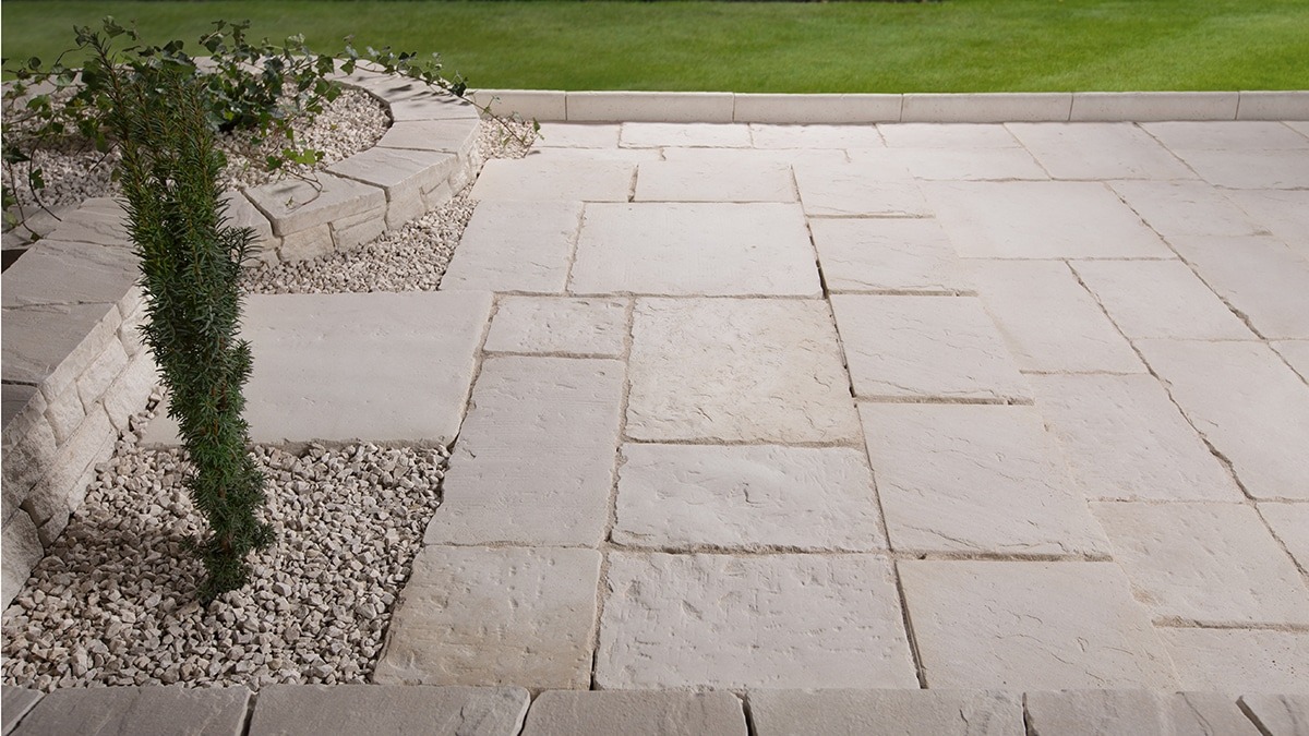 Jasnoszara, nieregularna nawierzchnia z kamiennych płyt, obrzeża z kostki, rabata z iglakiem i białym grysem na tle trawnika.