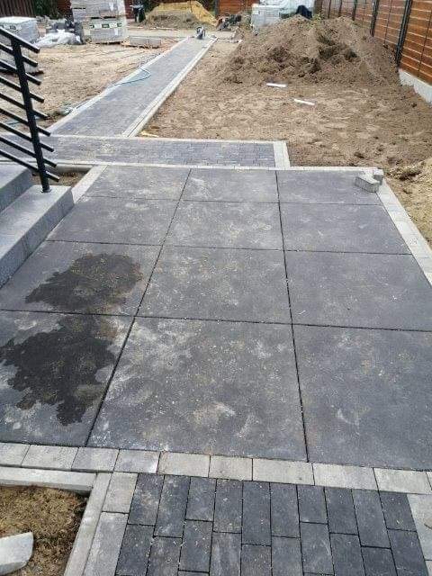 Fragment tarasu wyłożonego szarą kostką brukową, obrzeża z jasnoszarej kostki, w tle ścieżka z kostki prowadząca wzdłuż ogrodzenia, widoczny też stos kostki brukowej i pryzma ziemi.