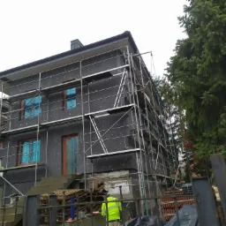 Budynek w trakcie ocieplania, zabezpieczone okna folią, widoczne rusztowania i pracownik w odblaskowej kamizelce na tle zielonych drzew.