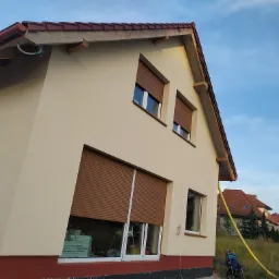 Jasna elewacja domu jednorodzinnego z brązowymi roletami okiennymi, widoczna rura doprowadzająca materiał elewacyjny oraz fragment niezabezpieczonego dachu z widocznymi krokwiami.