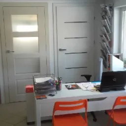 Wnętrze biura z dwoma białymi drzwiami: jedne z matową szybą, drugie z poziomymi paskami. Na pierwszym planie białe biurko z pomarańczowymi krzesłami, laptopem i materiałami biurowymi.