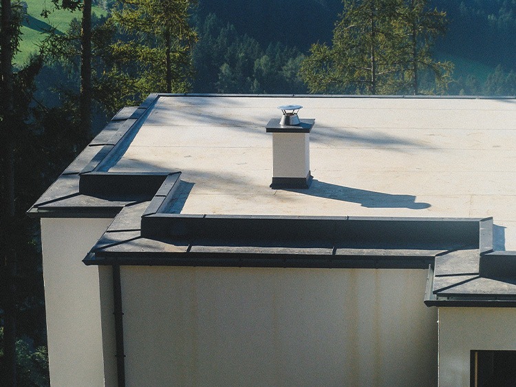 Płaski dach budynku z wykończeniem z ciemnej blachy, widoczny komin z metalową nasadą, tło stanowią zielone drzewa.