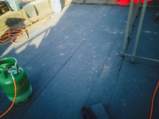 Zbliżenie na papę termozgrzewalną na dachu z widoczną butlą gazową i palnikiem dekarskim w trakcie pracy.