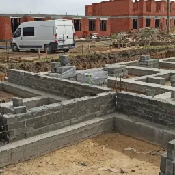 Wykonywanie fundamentów z bloczków betonowych na budowie, widoczne zbrojenia, w tle budynki w stanie surowym otwartym i biały bus.