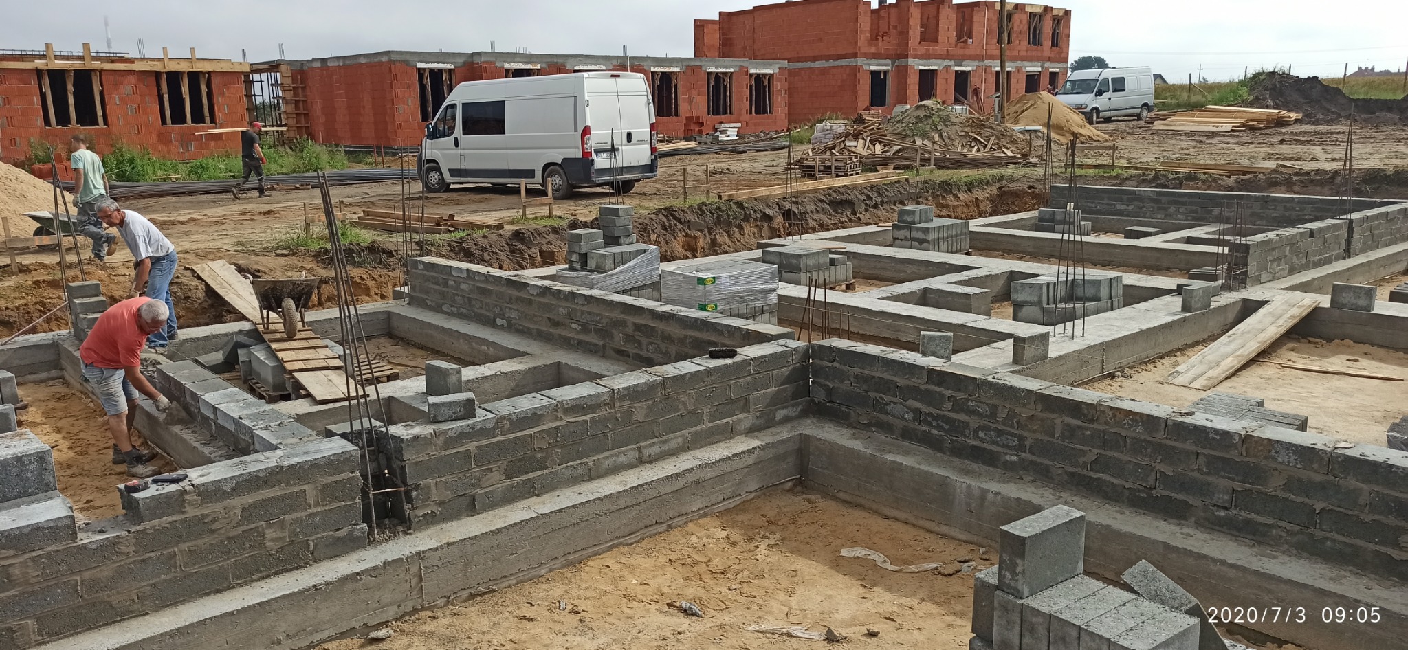 Wykonywanie fundamentów z bloczków betonowych na budowie, widoczne zbrojenia, w tle budynki w stanie surowym otwartym i biały bus.
