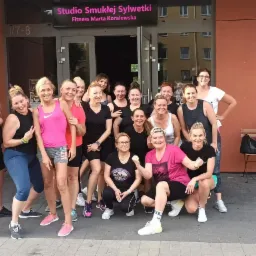 Grupa uśmiechniętych kobiet pozuje przed wejściem do studia fitness 'Studio Smukłej Sylwetki Fitness Marta Koralewska'. Niektóre kobiety ubrane są w sportowe stroje, inne w codzienne ubrania. W tle...
