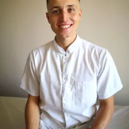 Uśmiechnięty młody mężczyzna w białym uniformie medycznym siedzi na białym łóżku rehabilitacyjnym, spoglądając w obiektyw.