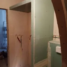 Konstrukcja z płyt gipsowo-kartonowych w trakcie montażu, widoczne przewody elektryczne wystające ze ściany, szpachlowane łączenia, metalowa drabina w tle.