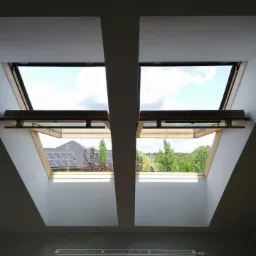 Dwa otwarte okna dachowe z drewnianą ramą, widok na zewnątrz z zielenią i domami z panelami słonecznymi, biała ściana i grzejnik poniżej.