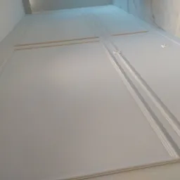 Ściana z zamontowaną płytą kartonowo-gipsową, pomalowana na biało, z dekoracyjnymi listwami przypodłogowymi i ornamentami w narożach.