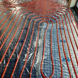 Montaż ogrzewania podłogowego: czerwone rurki zamocowane do siatki na folii izolacyjnej, widoczne białe zaciski mocujące.