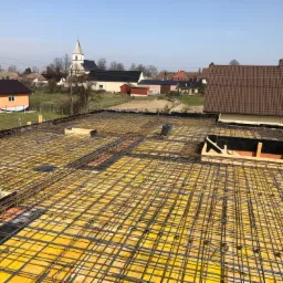 Zbrojenie płyty fundamentowej na żółtym szalunku, widok z góry na tle zabudowy wiejskiej z kościołem w oddali.