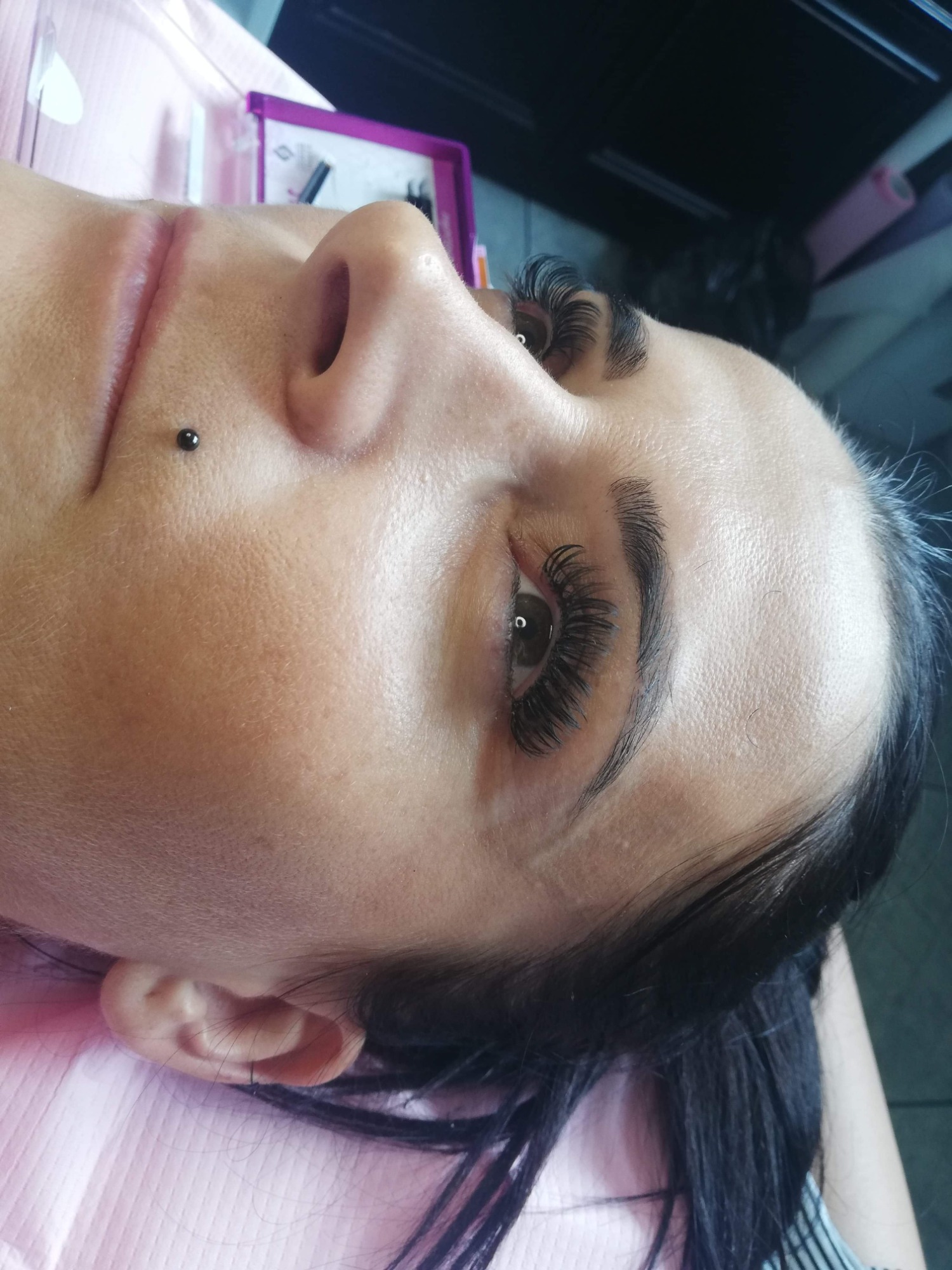 Kobieta leżąca na różowym podkładzie z nowymi, gęstymi, czarnymi rzęsami i delikatnym makijażem brwi, widoczny kolczyk na policzku.