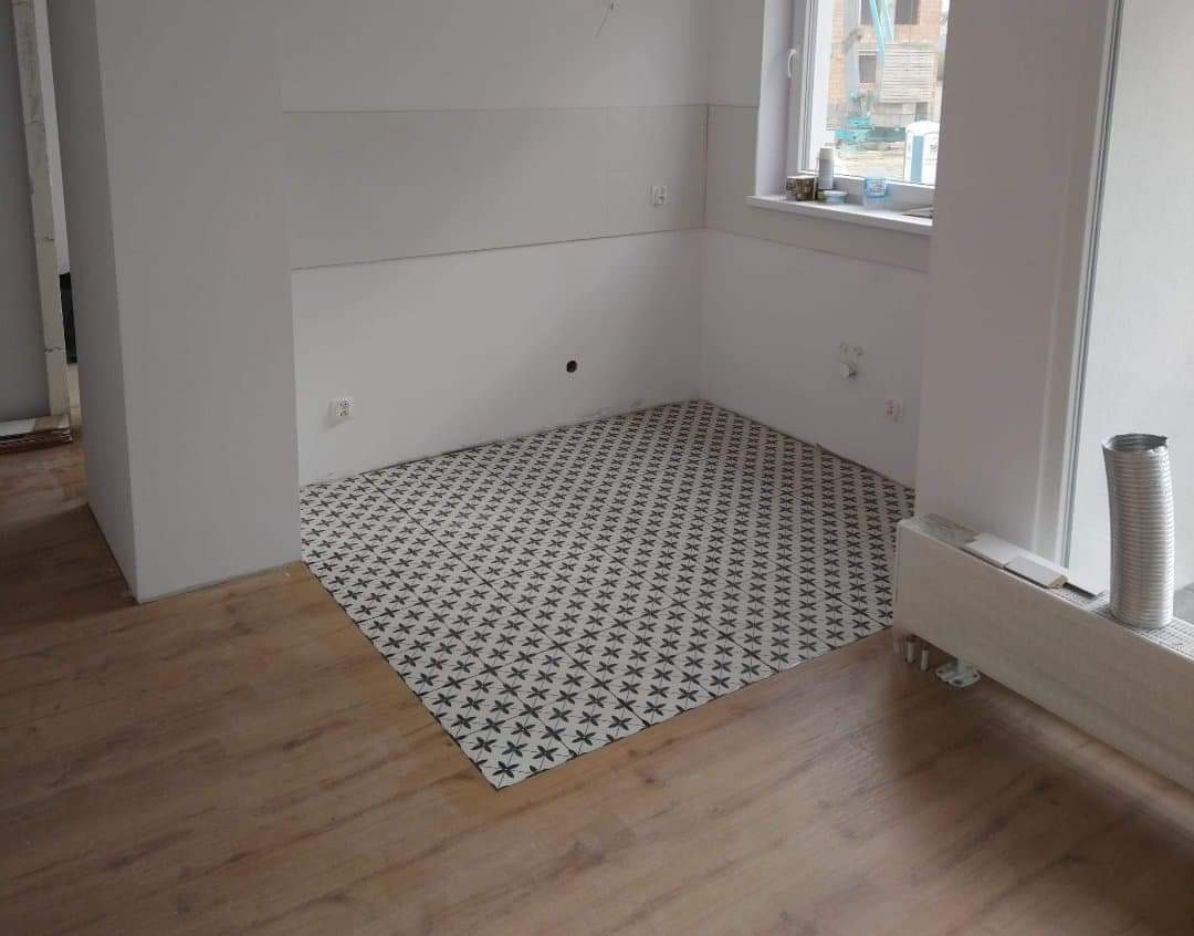 Fragment pomieszczenia w trakcie wykańczania: ułożona podłoga z płytek z wzorem gwiazdek na tle drewnopodobnych paneli, ściany przygotowane do dalszych prac wykończeniowych, widok na budowę przez...