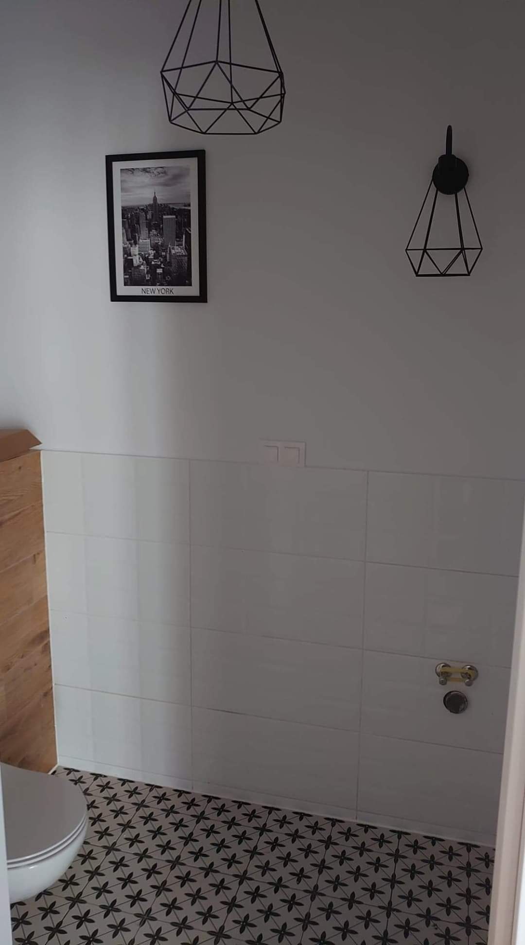 Wykończona łazienka w stylu minimalistycznym z białymi płytkami ściennymi, wzorzystą podłogą, drewnianym panelem bocznym, geometrycznymi lampami sufitowymi i czarno-białym plakatem z widokiem...