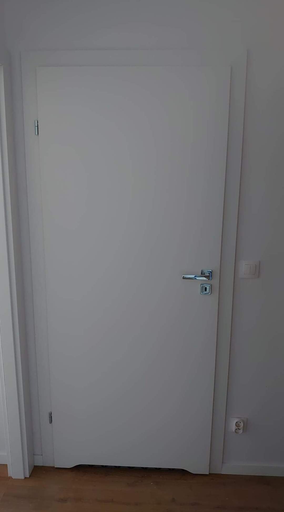 Zamknięte, pomalowane na biało drzwi wewnętrzne z chromowaną klamką i zamkiem, osadzone w białej futrynie, widoczny fragment podłogi i gniazdko elektryczne.