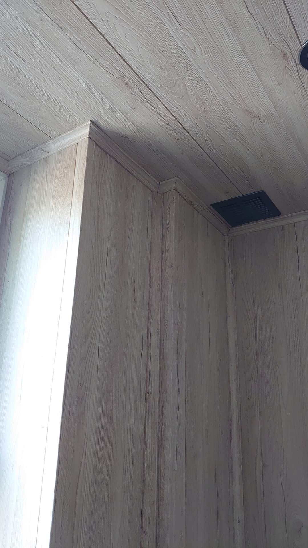 Ściana i sufit wykończone panelami imitującymi drewno, widoczny narożnik i kratka wentylacyjna.
