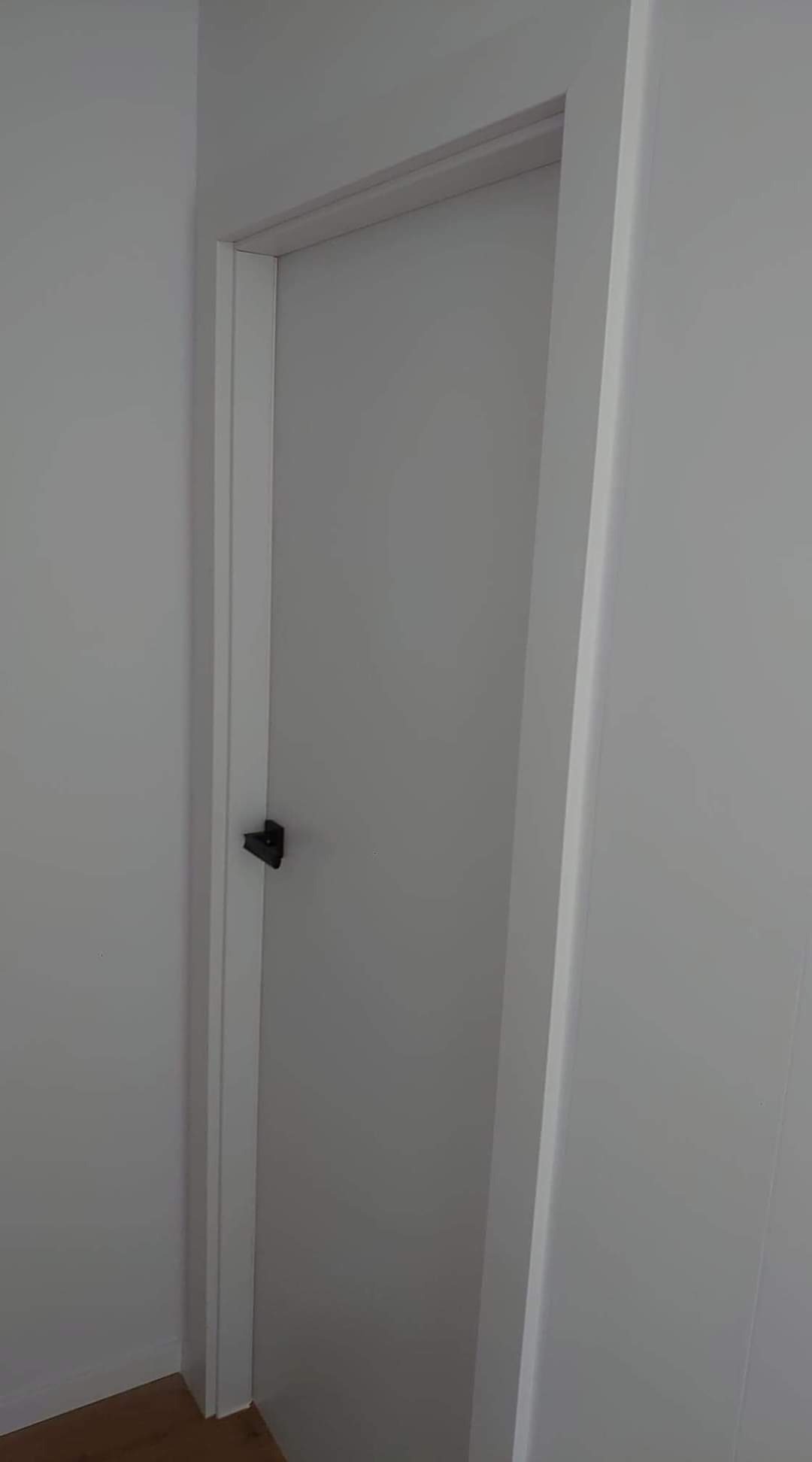 Zamknięte białe drzwi z czarną klamką w minimalistycznym wnętrzu, widoczne fragmenty białych ścian i ościeżnicy.