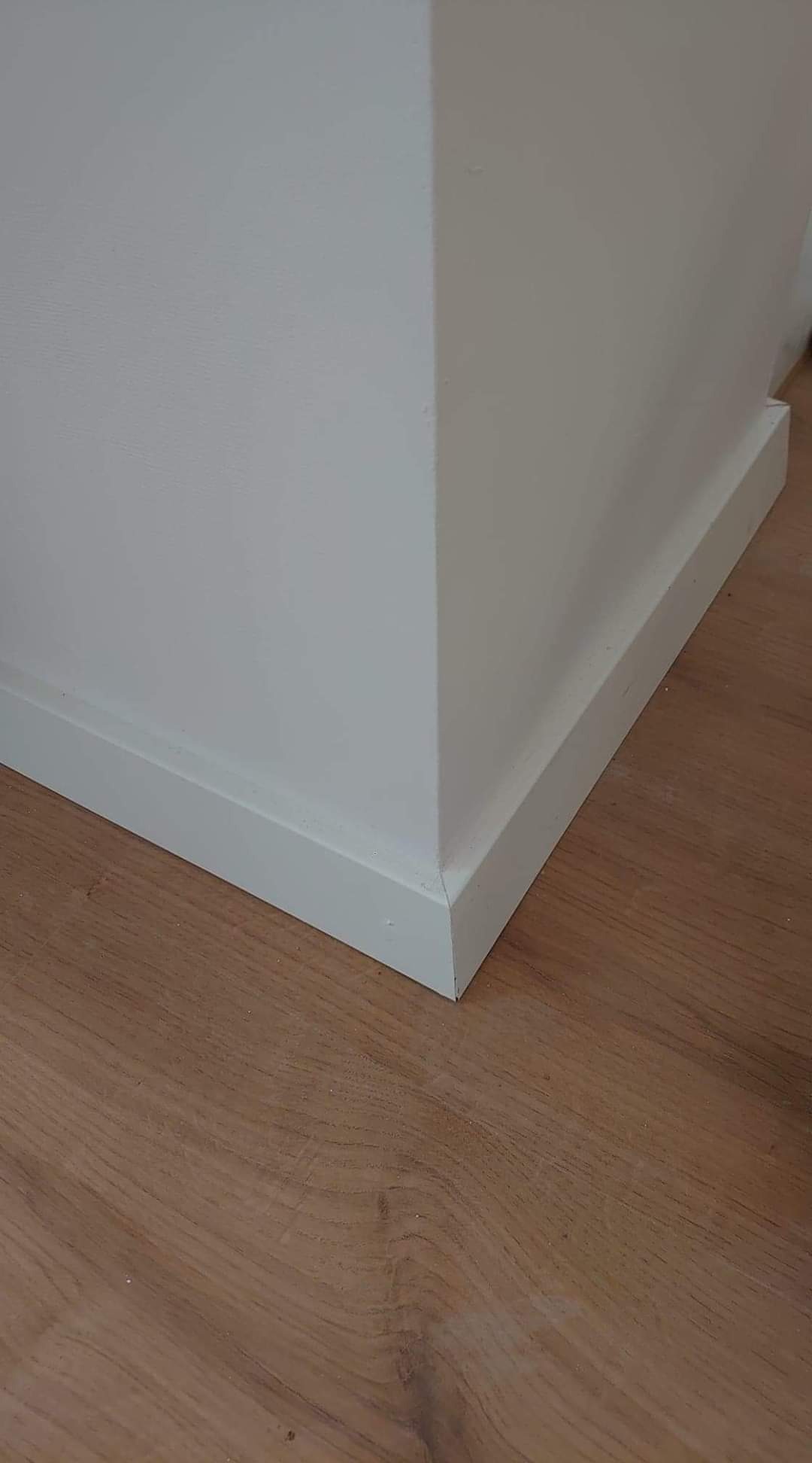 Pomieszczenie po remoncie: Ściana pomalowana na biało z zamontowaną białą listwą przypodłogową i brązowymi panelami podłogowymi.