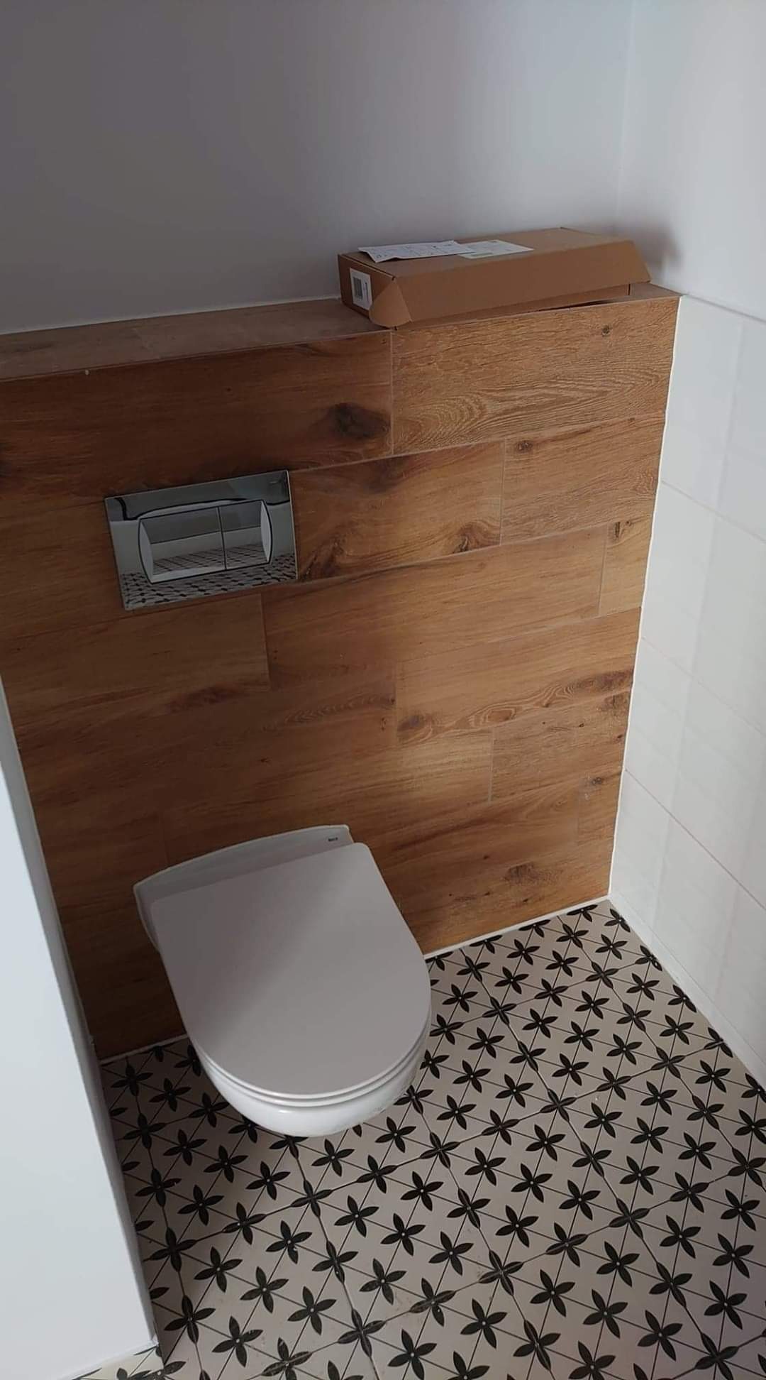 Zabudowa stelaża WC z drewnopodobnych paneli ściennych, chromowany przycisk spłuczki, biała miska ustępowa, podłoga z płytek w kwiatowy wzór, karton na górze zabudowy.