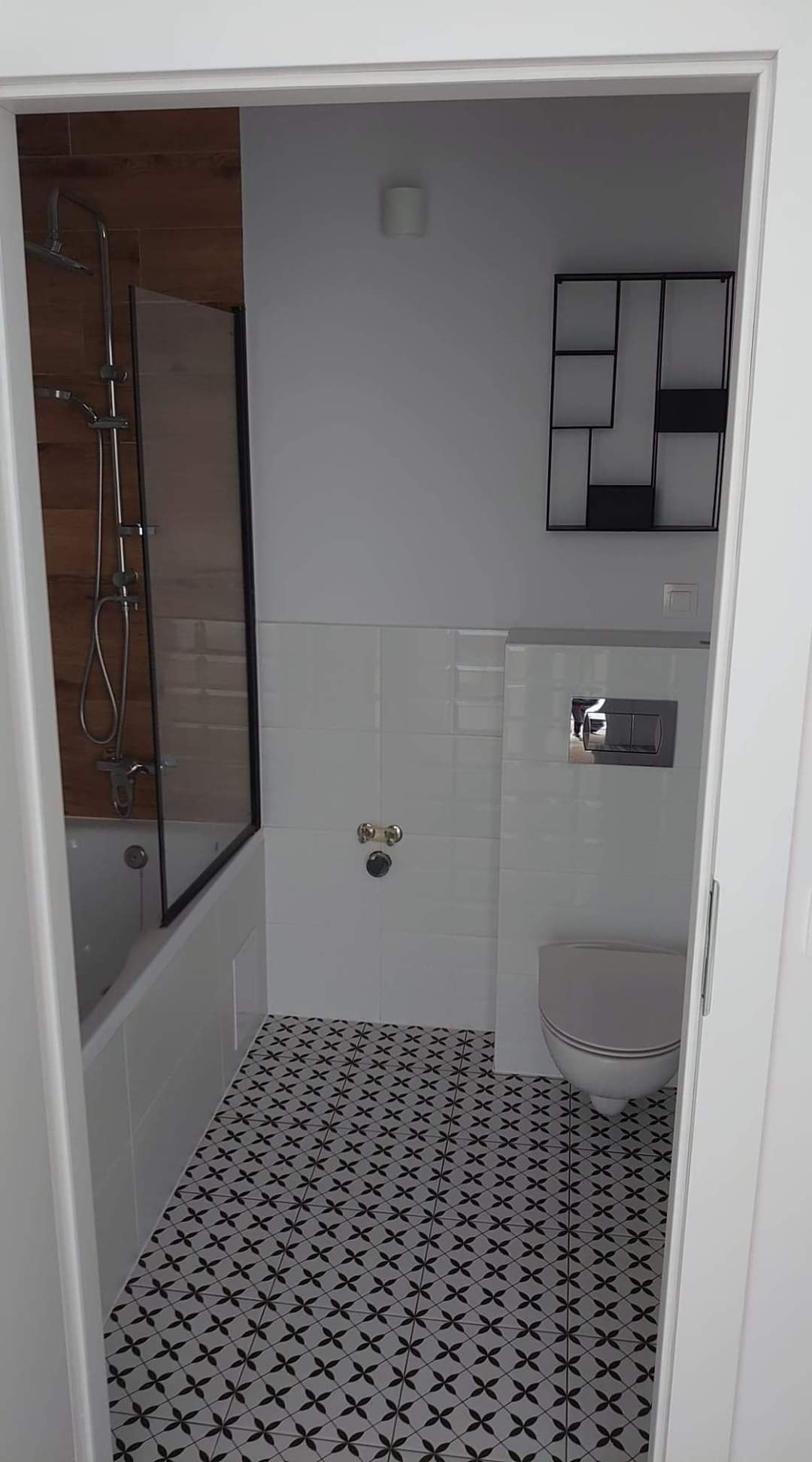 Wykończona łazienka z wanną z parawanem, białą glazurą, czarno-białą podłogą w gwiazdki i wiszącą, minimalistyczną półką na ścianie.