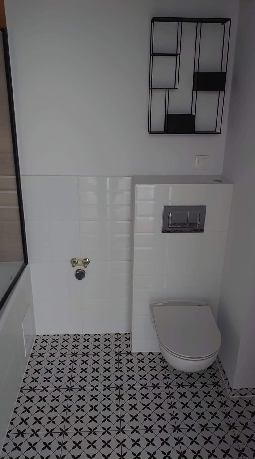 Wykończona łazienka z białymi płytkami ściennymi, toaletą podwieszaną z chromowanym przyciskiem spłukiwania oraz czarno-białą podłogą w geometryczny wzór, uzupełniona czarną, metalową półką...