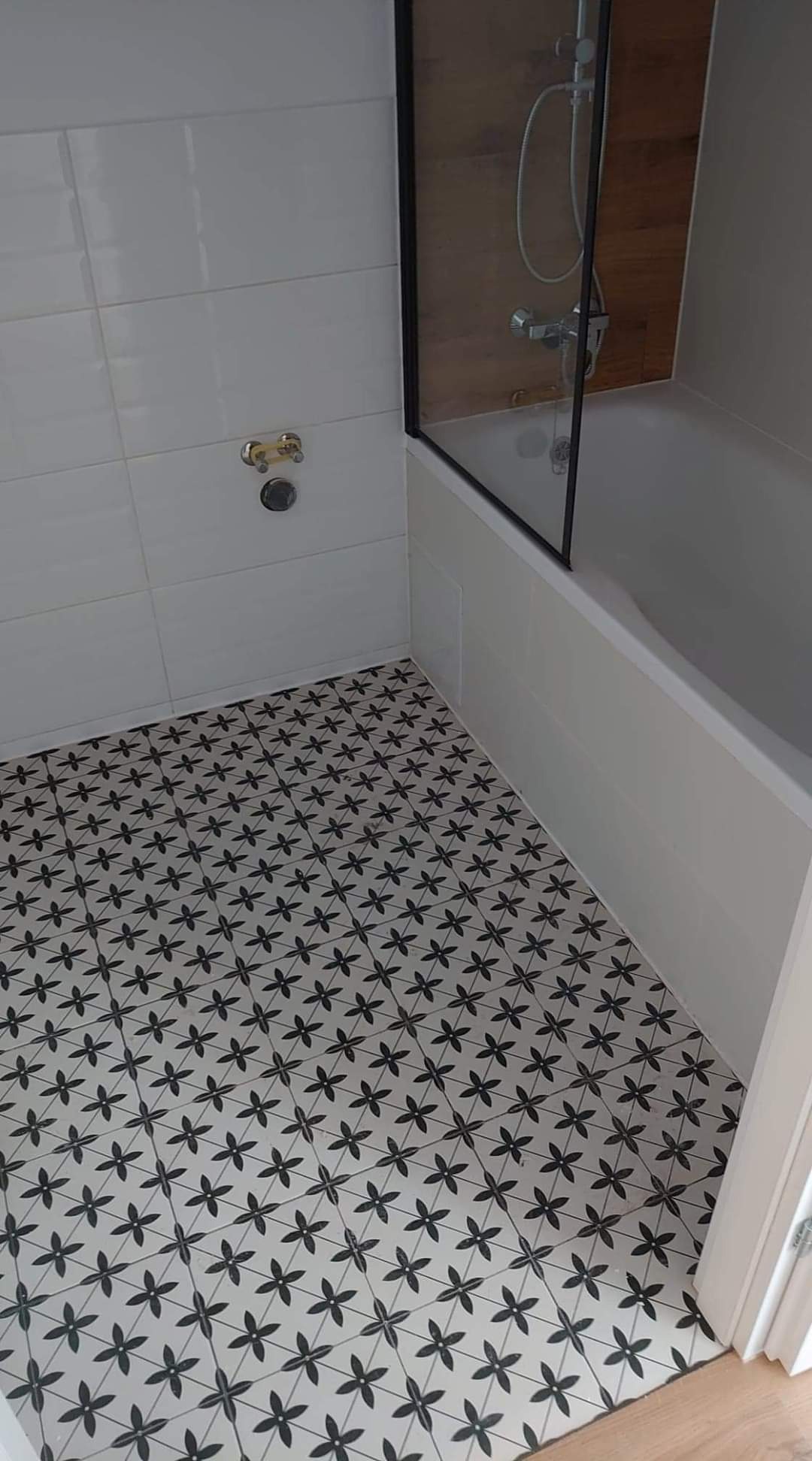 Biała łazienka z wanną za szklanym parawanem i podłogą wyłożoną wzorzystymi, czarno-białymi płytkami.