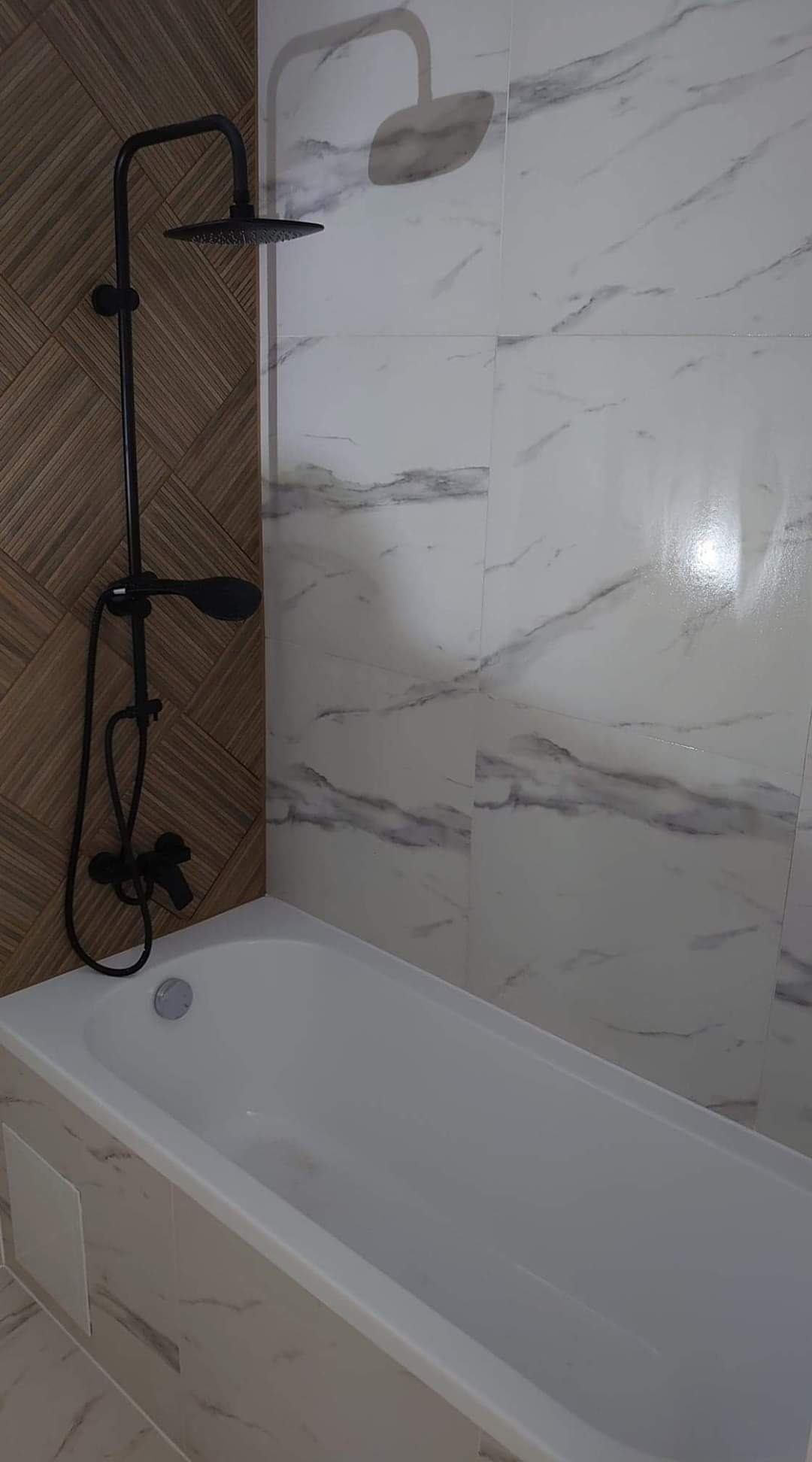 Nowoczesna łazienka z białą wanną, czarną armaturą prysznicową na tle drewnopodobnych i marmurkowych płytek ściennych.