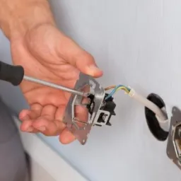 Elektryk przykręca gniazdko elektryczne do ściany za pomocą śrubokręta z żółto-czarną rączką. Widoczne przewody i puszka podtynkowa.