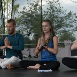 Cztery osoby siedzą w pozycji lotosu na scenie podczas plenerowej sesji relaksacyjnej, kobieta z warkoczykami prowadzi ćwiczenie z dłońmi złożonymi w geście namaste.