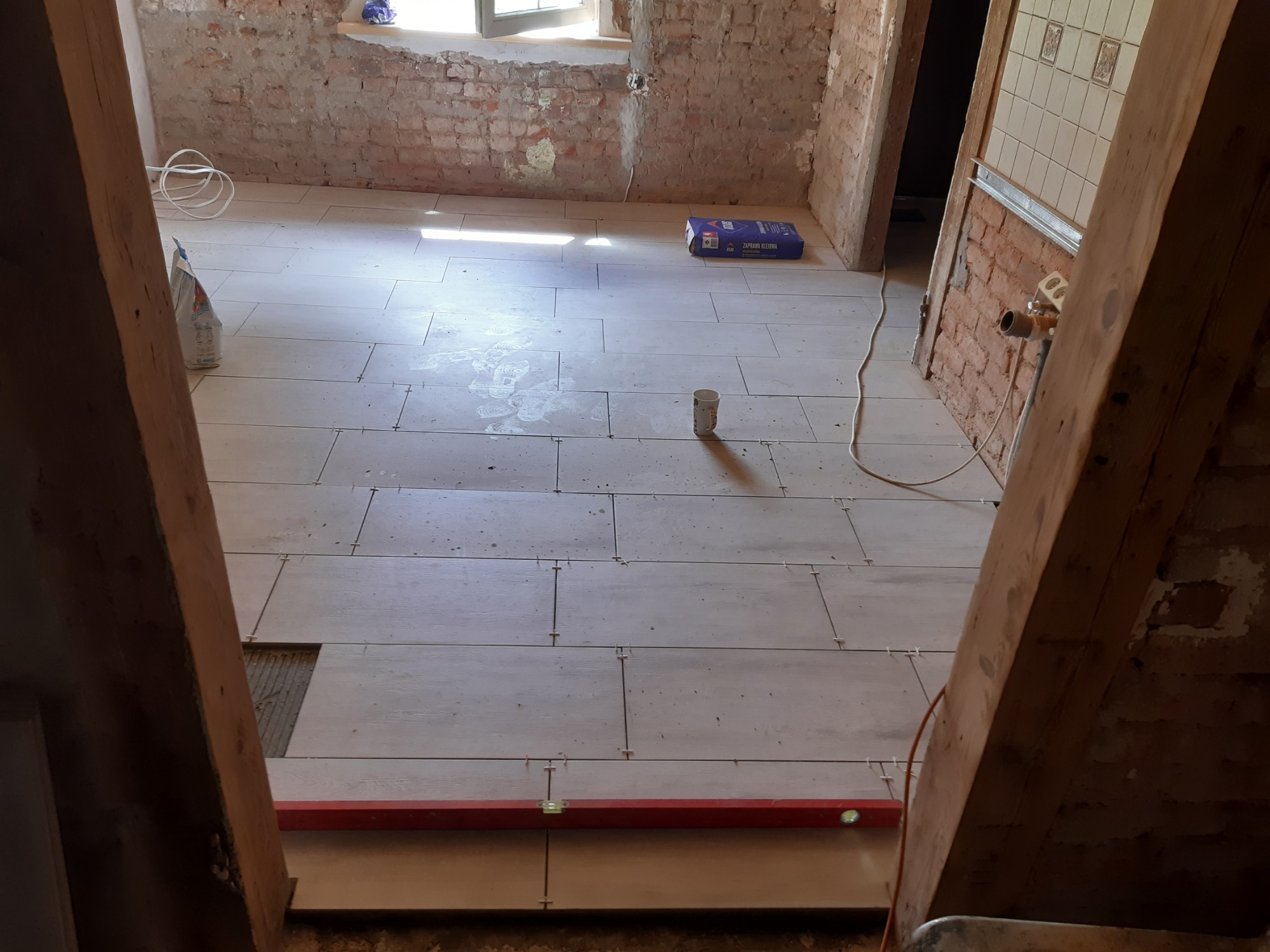 Układanie podłogi z płytek imitujących drewno w surowym wnętrzu, widoczny poziomica, krzyżyki dystansowe i odsłonięta cegła na ścianach.