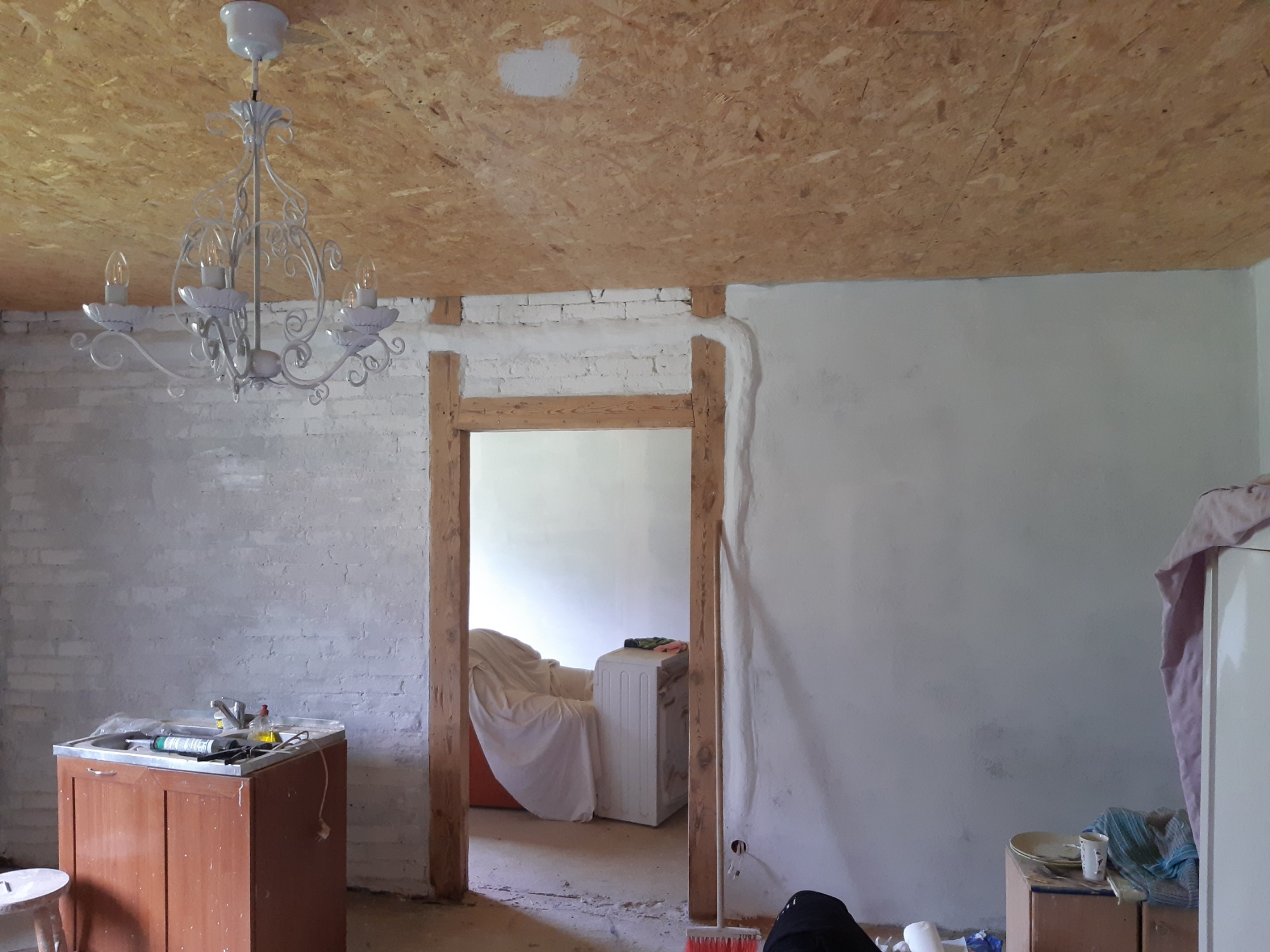 Wnętrze w trakcie remontu: surowy strop z płyt OSB, otynkowana ściana z fragmentami cegieł, drewniane obramowanie drzwi, widoczny zlew z narzędziami i biały żyrandol.