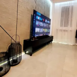 Nowoczesny salon z telewizorem na czarnej szafce RTV, dekoracyjną ścianą w beżowych tonacjach i dużym oknem z białymi zasłonami. Uwagę przyciągają detale dekoracyjne.