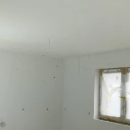 Wnętrze pomieszczenia po świeżo wykonanych pracach wykończeniowych, widoczne gładkie, białe ściany, okno zabezpieczone folią, wystające przewody elektryczne i lampa halogenowa na statywie...