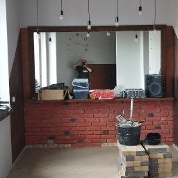 Szymon Szymański - Ściana z cegły klinkierowej wewnątrz pomieszczenia, nad nią lustro z drewnianą ramą i wiszące żarówki, na podłodze płytki, widoczne narzędzia i materiały budowlane, w lustrze odbija się osoba...